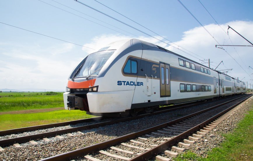 A double-decker Azerbaijan Railways passenger train travels on an electrified railway line.