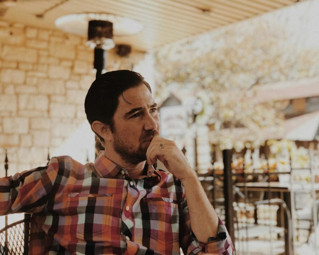 A man in a checked shirt sits indoors with his hand resting against his chin. Illustrative photograph.