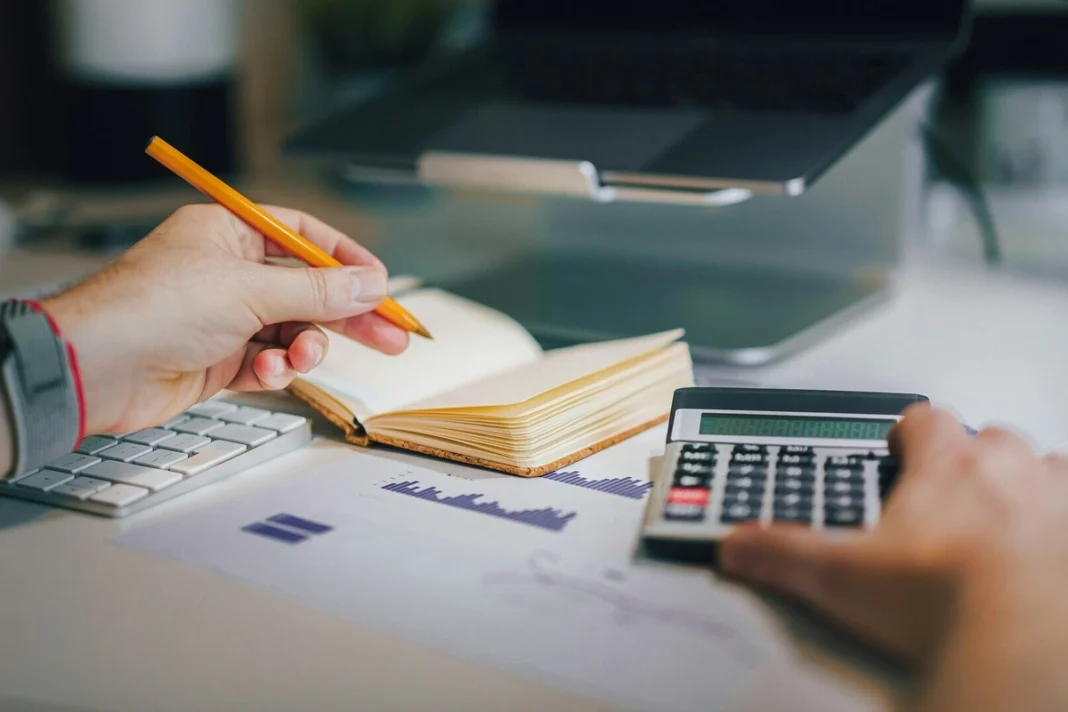 Person using a calculator and taking notes beside a laptop